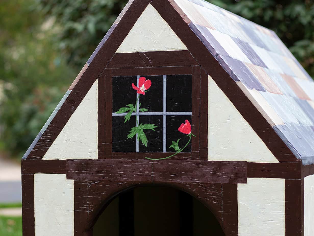 painted dog house with red flowers hanging behind a window